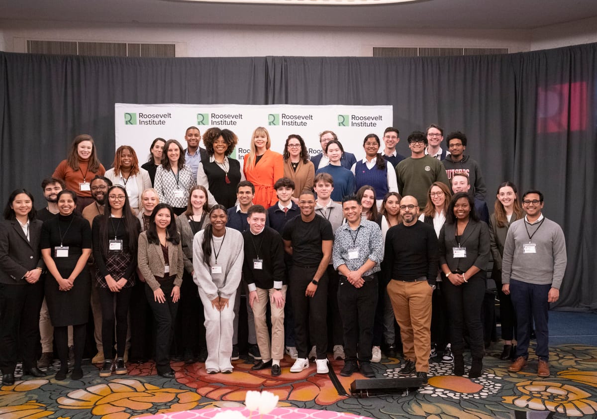 A diverse group of people pose together in front of Roosevelt Institute banners, smiling for a group photo in a conference room with gray curtains and a colorful carpet.