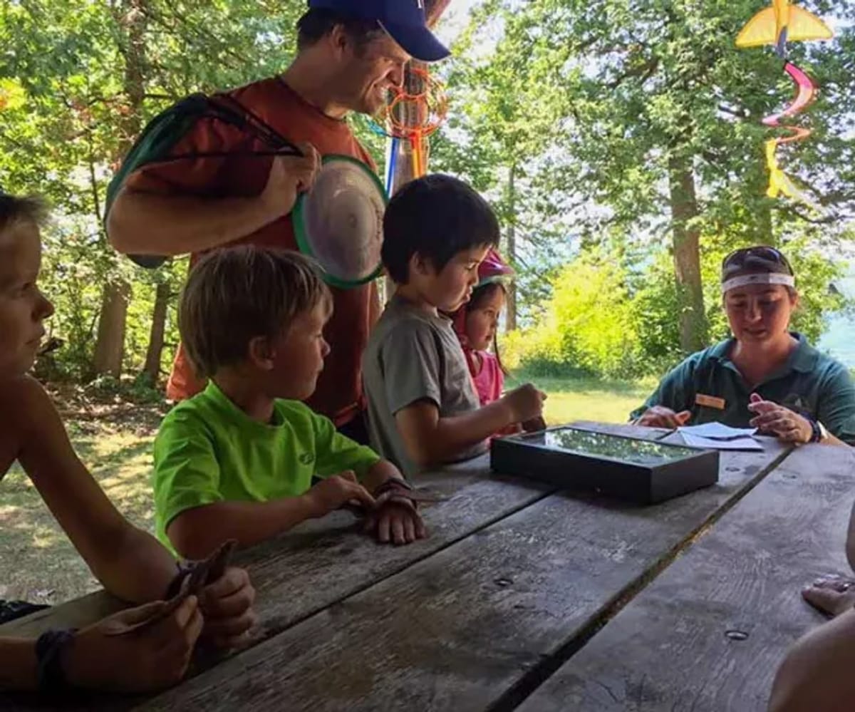A man smiling while standing next to his family at a picnic table where a naturalist is talking at a Vermont State Park.